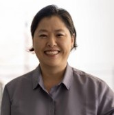 A middle aged Asian woman with medium length dark hair and a bright smile. She is wearing a light brownish grey shirt,